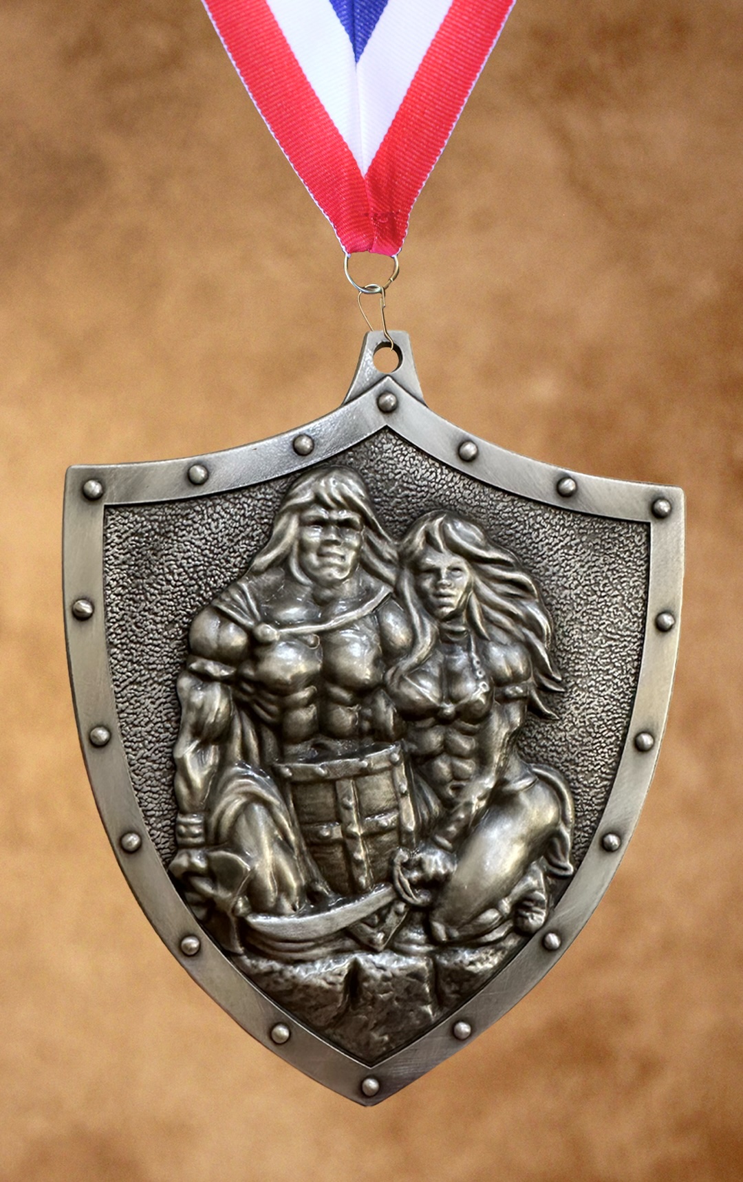 platinum pewter colored shield-shaped medal hanging from a red, white, and blue striped ribbon, featuring an embossed relief of a muscular warrior couple in heroic poses
